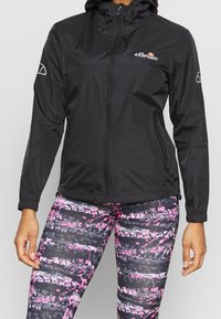 Svart lättviktsjacka med dragkedja framtill, justerbar huva och elastiska muddar. Matchas med mönstrade leggings med rosa och grå ränder.