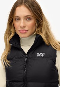 Schwarze gepolsterte Weste mit hohem Kragen, Reißverschluss und „SUPERDRY“-Logo auf der linken Brust. Über einem hellen Rollkragenpullover getragen.