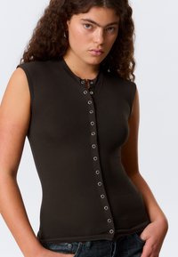 Tett, ermeløs topp i mørkebrun bomullsblanding med rund hals og dekorative sølvfargede trykknapper nedover fronten.