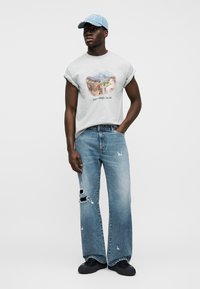 Muž oblečený v svetlosivej tielkovej grafickej tričku, svetlomodrých roztrhaných dlhých širokých džínsoch, čiernych topánkach a svetlomodom šiltovke, stojaci s jednou rukou v vrecku.