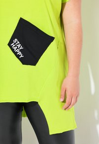 Camiseta verde lima con un bolsillo negro que lleva el texto "MANTENTE FELIZ", combinada con leggings negros. Tela suave, corte relajado y dobladillo curvado.