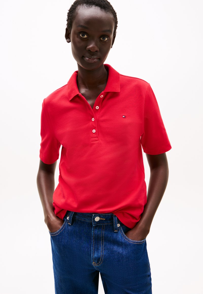 Rode polo met een kraag, voorzien van een knoopsluiting met drie knopen en een klein logo. Gecombineerd met donkerblauwe jeans.