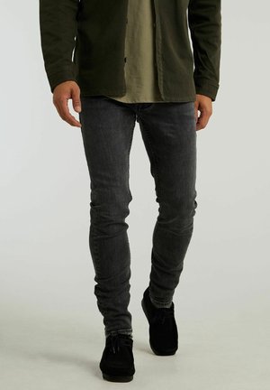 Homme portant un jean skinny noir délavé, des chaussures noires, une chemise vert olive et une veste vert foncé marchant sur un fond gris clair.