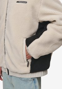 Giacca in pile con sezioni crema e nere, dotata di tasca con zip, polsi elastici e bordo ribbed. Texture morbida in tutto il capo.