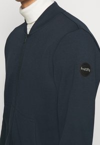 Felpa con zip blu navy con collo alla coreana, dotata di tasche frontali e una toppa rotonda con logo nero sulla manica sinistra. Tessuto dalla trama morbida.