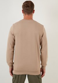 Sudadera beige de manga larga, con cuello acanalado y una superficie texturizada. La vista posterior muestra un corte relajado y un dobladillo recto.