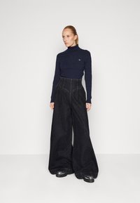Maglione a collo alto a coste di colore navy abbinato a pantaloni in denim neri a vita alta con gamba larga. Le scarpe presentano una suola nera spessa con accenti metallici.