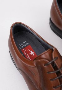 Zapatos de cuero marrón con una textura suave, cierre de cordones y una suela de goma negra. El interior cuenta con una etiqueta de marca en rojo y gris.