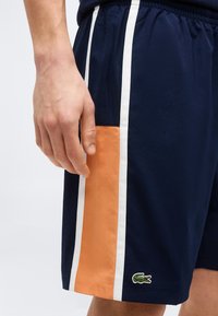 Κοντινό ενός χεριού που ακουμπάει δίπλα σε σορτς ναυτικού μπλε με κάθετες λευκές και πορτοκαλί ρίγες και πράσινο λογότυπο κροκοδείλου κοντά στο τελείωμα.