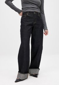 Jeans negros de pierna ancha con dobladillo gris en contraste, costuras doradas y diseño clásico de cinco bolsillos. Tela de mezclilla suave, ajuste relajado.