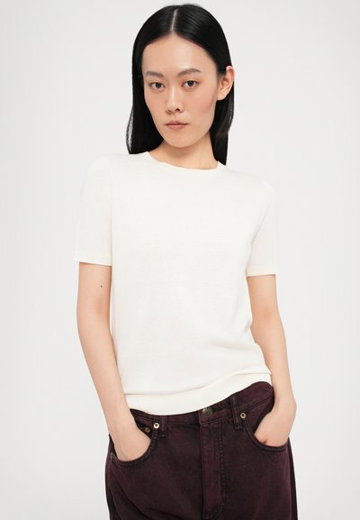 Maglione bianco a maniche corte con scollo rotondo, realizzato in un materiale morbido, abbinato a pantaloni bordeaux scuro con una texture leggermente consumata.