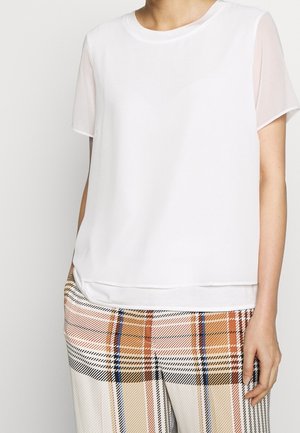 Frau trägt eine schlichte weiße Bluse mit kurzen Ärmeln, kombiniert mit hoch taillierter karierter Hose in Beige-, Orange-, Blau- und Schwarztönen.