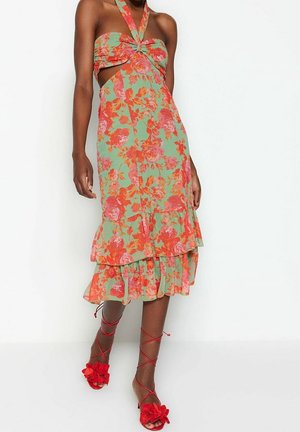 Blomstret midi-kjole i grøn med rosa og orange roser. Halterneck-design, åbne sideudskæringer, lagdelt flæsekant. Røde sandaler med stropper.