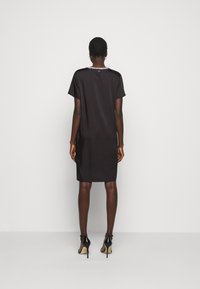 Černé šaty krátkého rukávu s rovnýcm střihem, hladkou texturou a dekorativním detailem na výstřihu. Nosí se s černými vysokými podpatky.