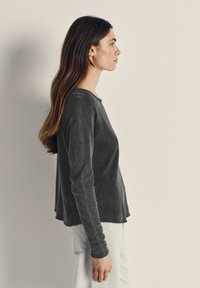 Pitkät ruskeat hiukset omaava nainen, jolla on päällään tummanharmaa pitkähihainen paita ja vaaleansiniset farkut, seisoo sivuprofiilissa yksinkertaisen beigen taustan edessä.