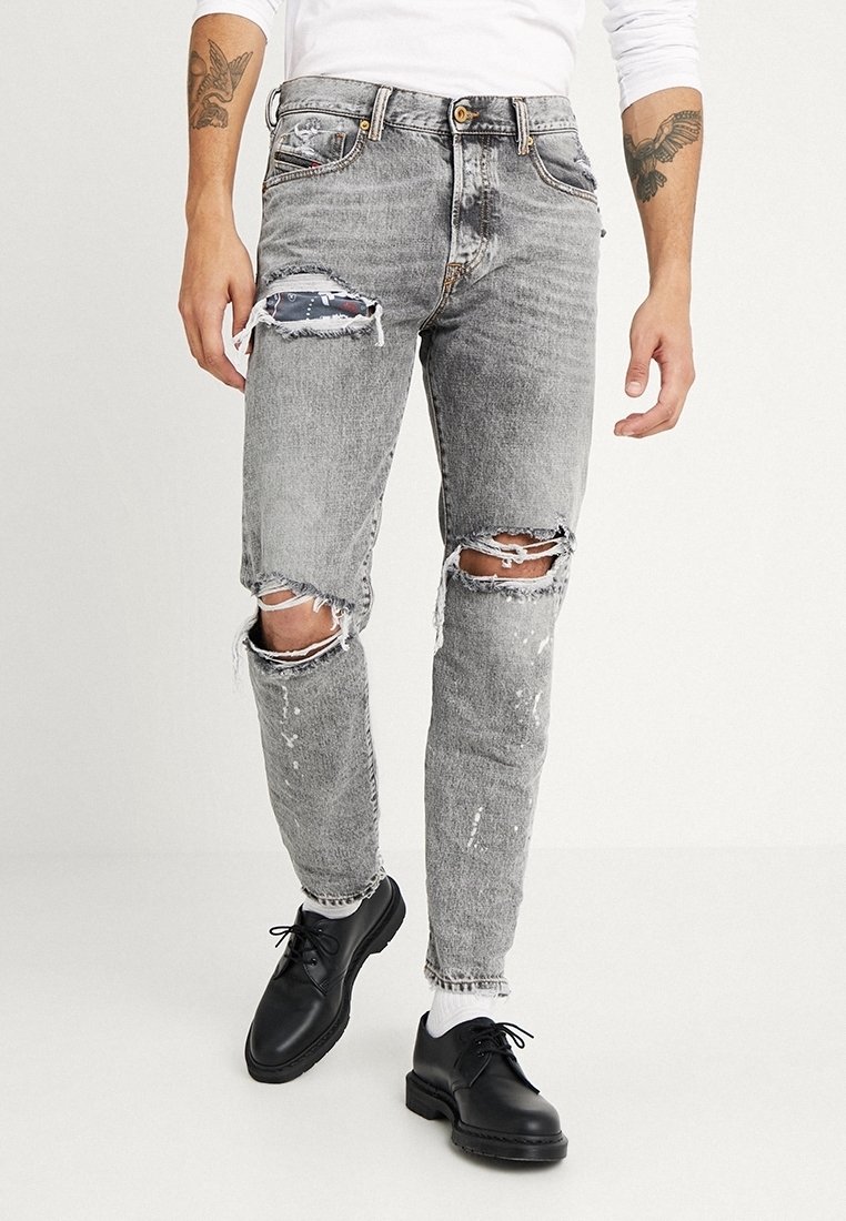 Person som bär grå slitna jeans med hål vid låren och knäna, svarta skor, vita strumpor och vit långärmad tröja, står på vit golv.
