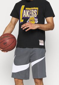 Maglietta nera con grafica viola e gialla dei "Los Angeles Lakers", abbinata a pantaloni corti grigi con design bianco del marchio Nike. Tiene in mano una palla da basket.