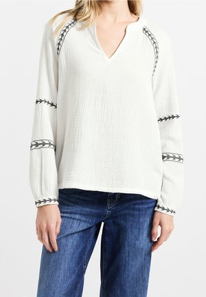 Kobieta ubrana w luźną białą bluzkę z długim rękawem, z czarnymi haftowanymi detalami na rękawach i w okolicy ramion, w połączeniu z niebieskimi dżinsami.