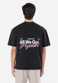 Tricou negru din bumbac cu un grafic mare pe spate, având textul "Only We Got. Pegador" în culorile alb și roz. Mâneci scurte, croi clasic.