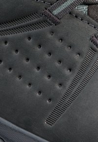 Svart sneaker med en texturerad mockaöverdel som har små perforeringar och randiga snördetaljer. Den har en gummisula och en slank design.