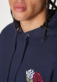 Sudadera azul marino con diseño de rosa bordado en rojo y blanco. Cuenta con cordones ajustables y ojales plateados. Textura de tela suave.