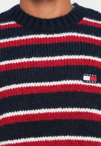 Jersey de marina con amplias rayas rojas y blancas. Cuello redondo acanalado. Presenta un pequeño parche de logo con "TOMMY JEANS" en azul marino, rojo y blanco.