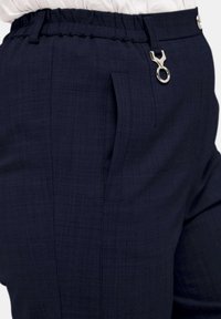Marineblaue Hose aus strukturiertem Stoff, mit elastischem Bund und einem silbernen Ring-Detail. Verfügt über seitliche Taschen für zusätzlichen Komfort.