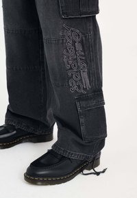 Pantalones cargo negros con diseño bordado, que cuentan con múltiples bolsillos, un corte holgado y una tela suave al tacto. Combinados con zapatos negros.