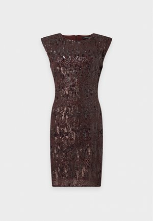 Un mannequin se tient devant un fond uni, vêtu d’une robe sans manches à paillettes brun foncé, de collants transparents et de sandales noires à talons hauts.