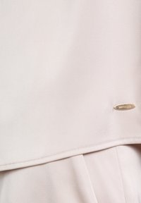 Helles rosa Bluse aus glattem Stoff mit umgeschlagenem Saum. Enthält ein kleines silbernes Logo-Tag mit dem Text "BETTY.CO." an der Seite.