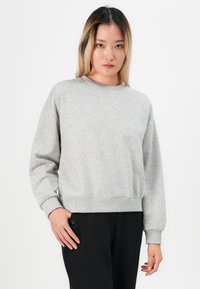 Ung kvinna med långt ombrehår bär en ljusgrå sweatshirt och svarta byxor, står med en hand nära fickan mot en vit bakgrund.