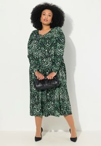 Vihreä ja musta kuvioitu pitkä mekko, jossa on pitkät hihat, pyöreä pääntie ja lentävä siluetti, yhdistetty mustaan laukkuun.