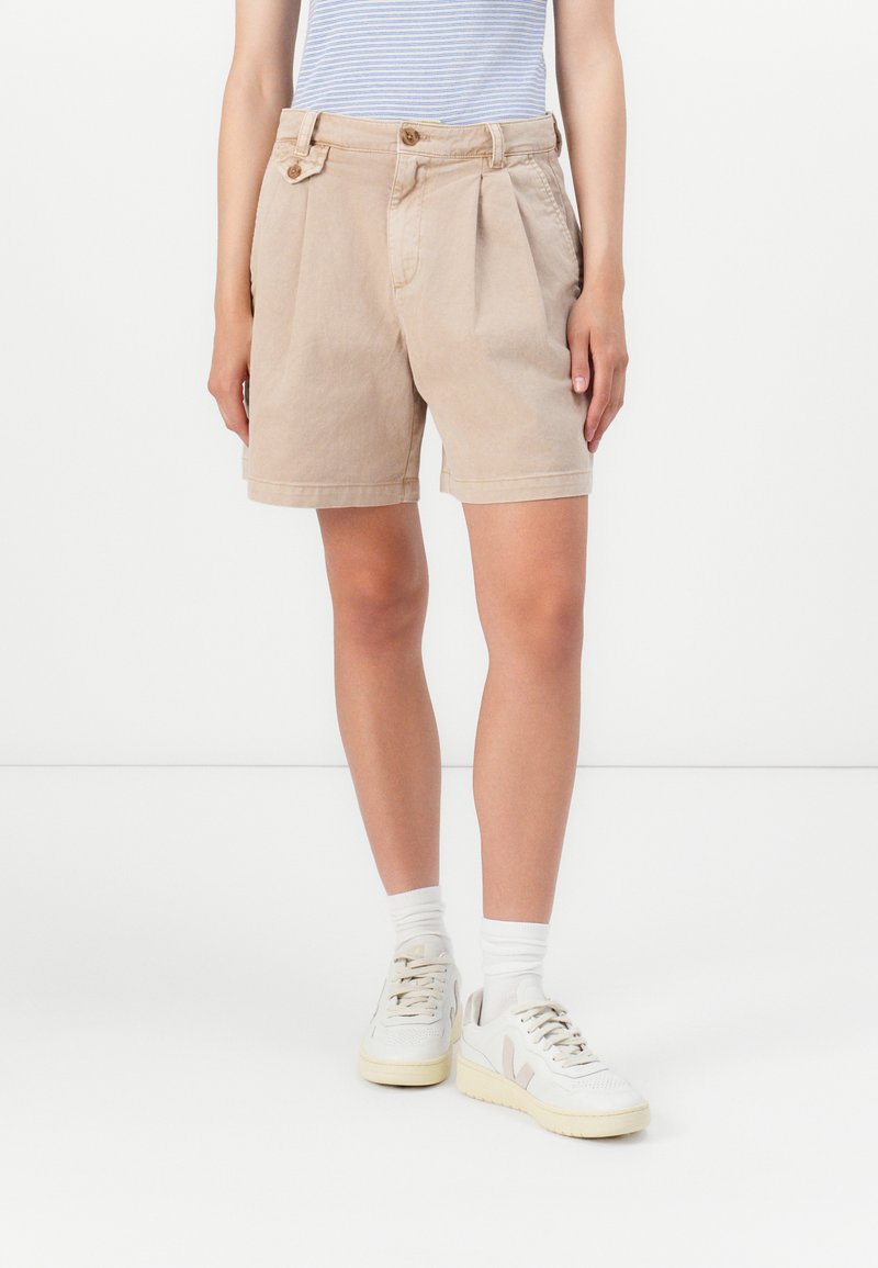 Shorts en coton beige avec plis, dotés d'une taille boutonnée et de poches latérales, associés à des baskets blanches et des chaussettes côtelées.