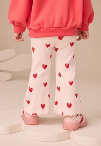 Svetloružové rebrované rozšírené nohavice s roztrúsenými červenými potlačami srdiečok, kombinované s ružovými teniskami. Mäkká látka s detailom volánikového lemu.