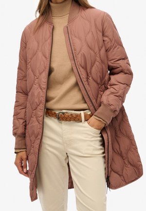 Femme portant une longue veste matelassée rose poudré sur un pull à col roulé beige et un pantalon crème avec une ceinture marron tressée.