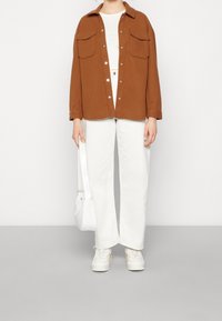 Brun skjorta med knappar och två bröstfickor, vit t-shirt, vita vida byxor, vita sneakers och liten vit axelväska.