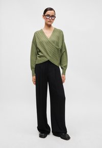 Πράσινο ζακέτα τύπου wrap με υφασμάτινες διαγώνιες ρίγες, συνδυασμένη με μαύρες παντελόνες φαρδιές και χοντρές μαύρες παπούτσια.