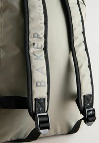 Sac à dos en tissu gris avec des bretelles noires, présentant un design rembourré et le mot "BAKER" en lettres en relief sur la sangle. Quincaillerie ajustable.