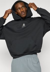 Μαύρο φούτερ με κουκούλα και μπροστινή τσέπη, με ανακλαστικό λογότυπο στο στήθος. Φορεμένο με γκρι φούτερ παντελόνι με ελαστική μέση.