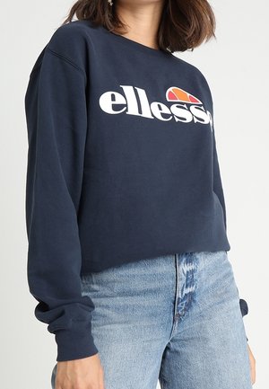 Femme portant un sweat-shirt ellesse bleu marine avec un logo blanc et un jean taille haute bleu clair, debout devant un fond uni.