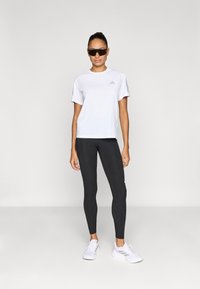 Weißes Sportshirt mit kurzen Ärmeln und Logo, kombiniert mit schwarzen Leggings und weißen Turnschuhen. Modell trägt dunkle Sonnenbrille und steht frontal zur Kamera.