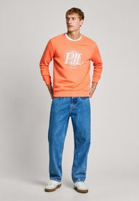 Sudadera naranja con logo y texto blanco, combinada con unos pantalones vaqueros azules holgados y zapatillas blancas. Presenta un cuello redondo y puños acanalados.