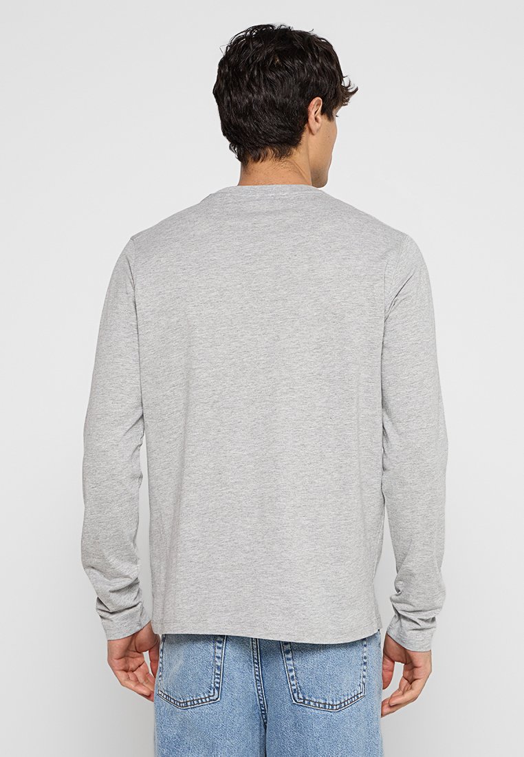 Mann in einem hellgrauen Langarmshirt und blauen Jeans, der mit dem Rücken zu einem schlichten weißen Hintergrund steht.