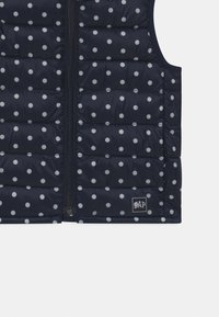 Námořnická modrá péřová vesta s bílými puntíky, která má zapínání na zip vpředu a malou logovou nášivku na spodním lemu. Hladká, prošívaná textura.