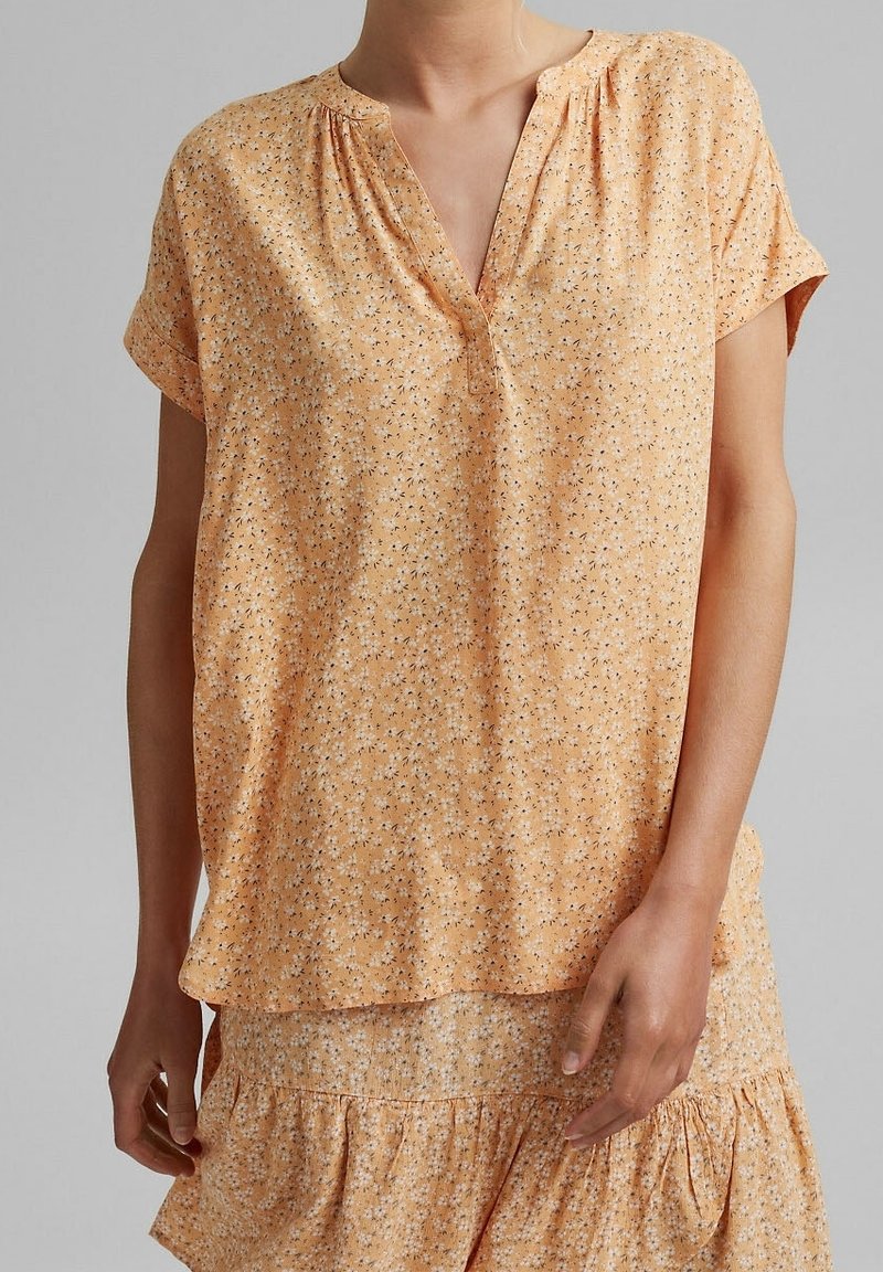 Kurze Ärmel, orangefarbener Top mit lockerer Passform, ausgestattet mit einem V-Ausschnitt und einem kleinen floralen Muster in Schwarz und Weiß. Weicher, strukturiertes Material.