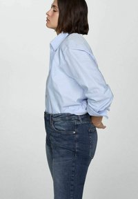 Svetlo modra srajca z gumbom in zavihanimi rokavi, v kombinaciji s temno modrimi ozkimi djeansami. Tkanine se zdijo gladke in prilegajoče.