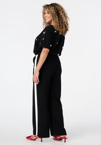 Femme aux cheveux bouclés, portant un pantalon noir avec une bande blanche, un haut noir à motifs blancs et des talons rouges, se tenant de profil sur fond blanc.
