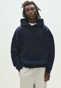 Navy blue oversized hoodie made of soft fabric with a kangaroo pocket and drawstring hood, paired with gray sweatpants.