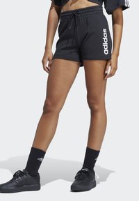 Čierne športové šortky s elastickým pásom, s bielym logom Adidas na boku, v kombinácii s čiernymi ponožkami a topánkami.