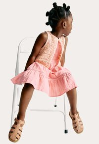 Robe rose avec un corsage texturé et une jupe évasée, assise sur une chaise, associée à des sandales à lanières marron clair. Profil d'un enfant visible.
